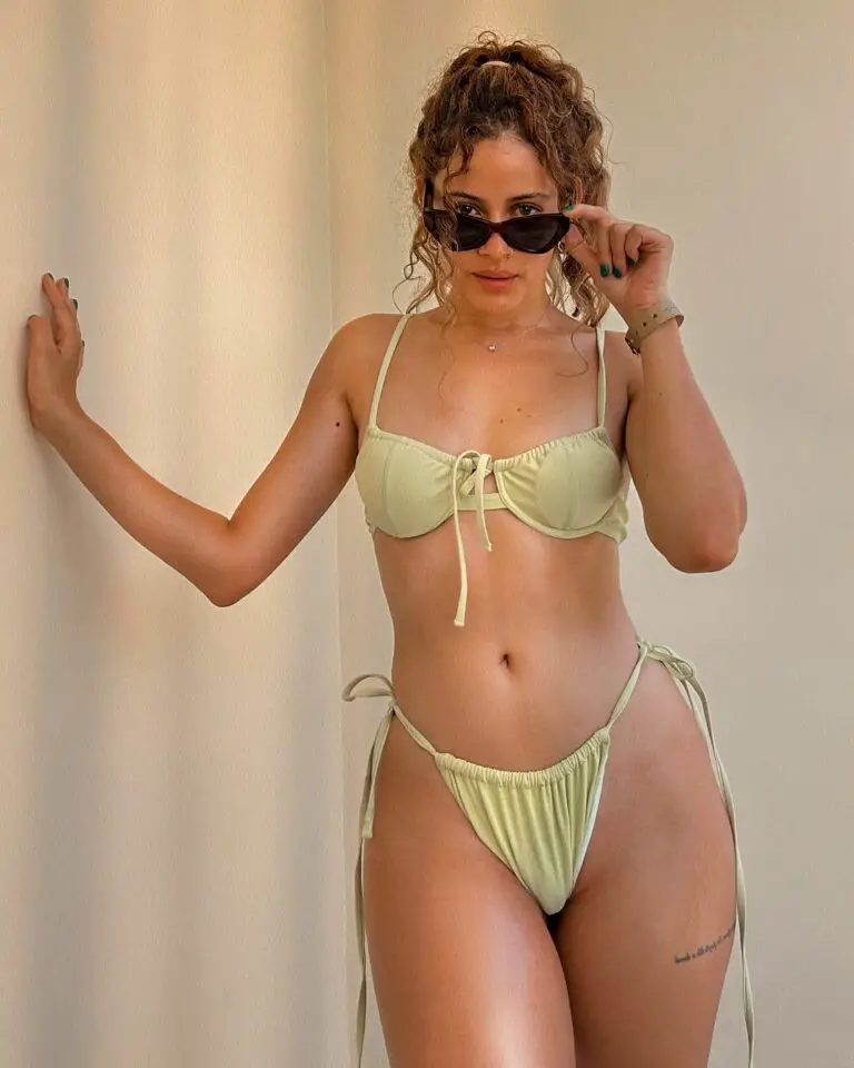 Alana in a pale green bikini, lifting her sunglasses while posing against a neutral wall.
